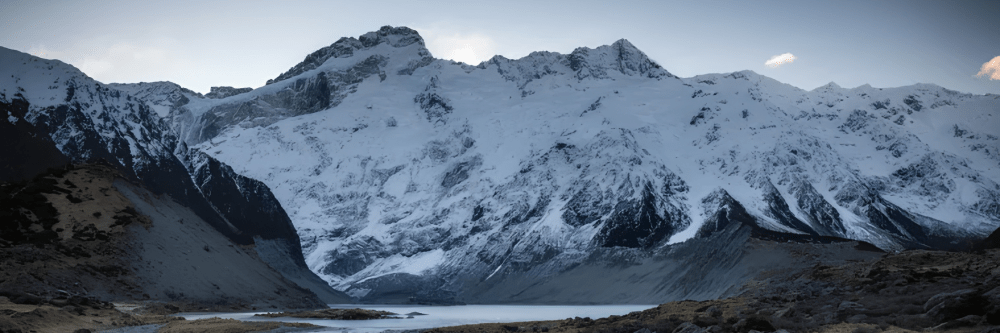 Snow-capped mountains with a lake and rocky foreground under a cloudy sky.