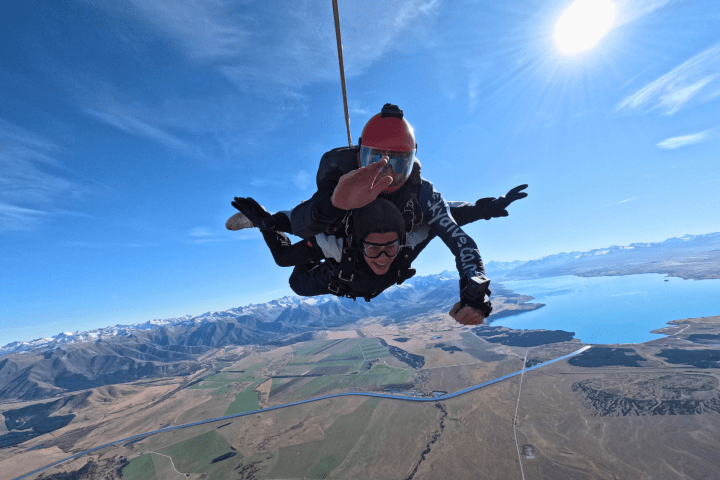 Two skydivers in free fall above scenic landscape with mountains, lake, and clear blue sky.