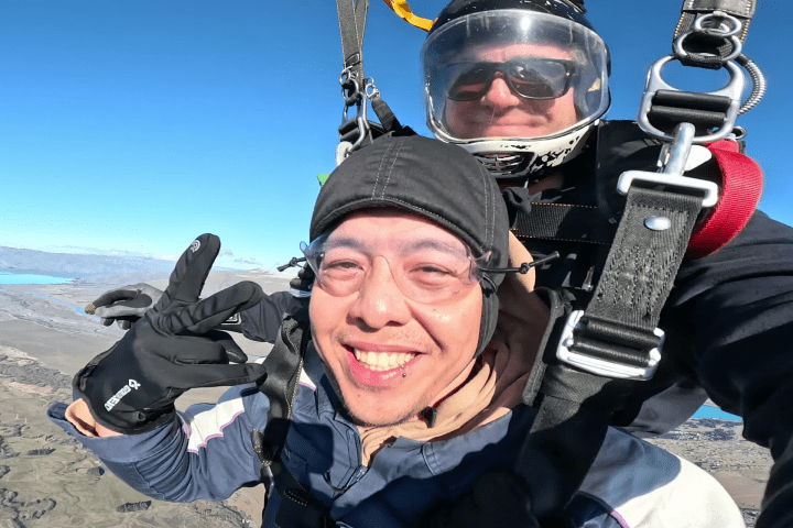 Two people tandem skydiving with parachute open, smiling faces, and clear blue sky in background.