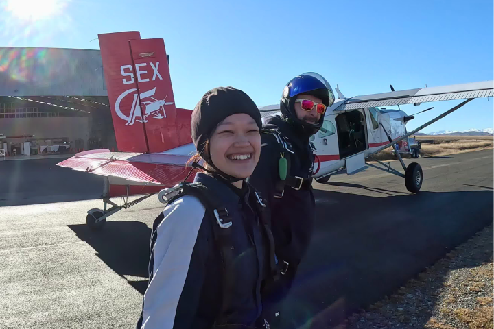 Two people in skydiving gear smiling in front of a small airplane on a sunny day.