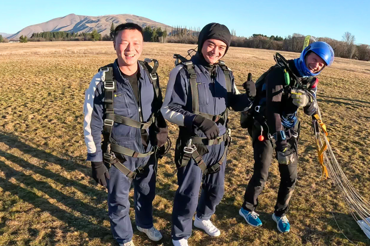 Three people in parachute gear smiling, standing on a grassy field.