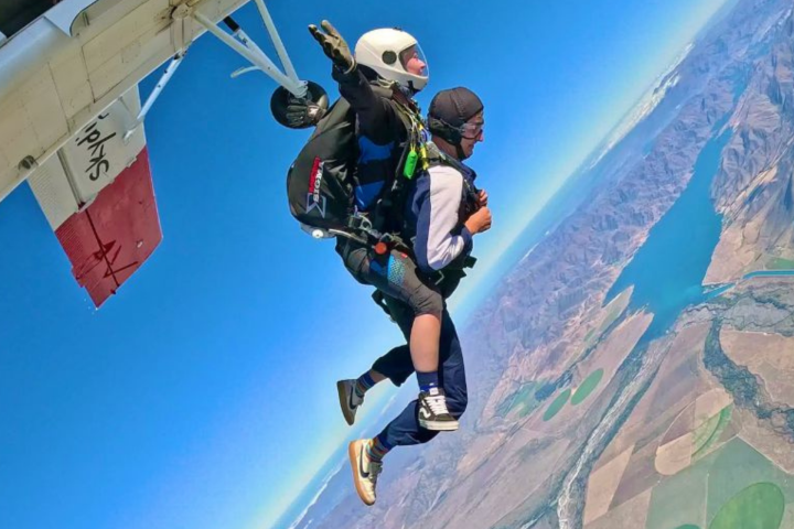 Two people tandem skydiving from a plane over a scenic landscape.