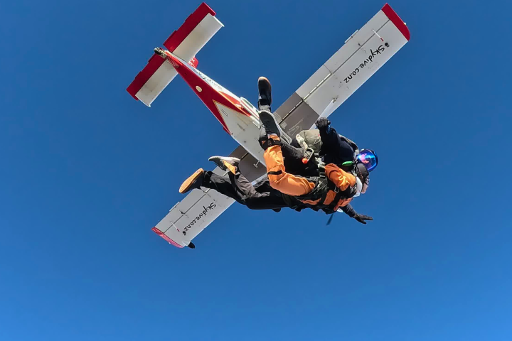 Two skydivers jump from an airplane against a clear blue sky.