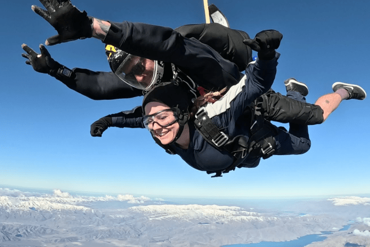 Two people tandem skydiving with mountains and a lake below.