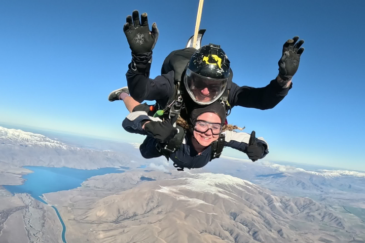 Two people tandem skydiving, smiling with thumbs up, over a landscape with mountains and water below.