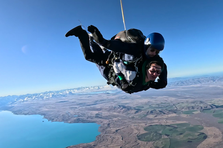Two people tandem skydiving above a scenic landscape with mountains and a lake.