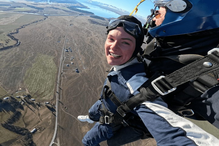 Person tandem skydiving with instructor, landscape below, both smiling and wearing helmets.