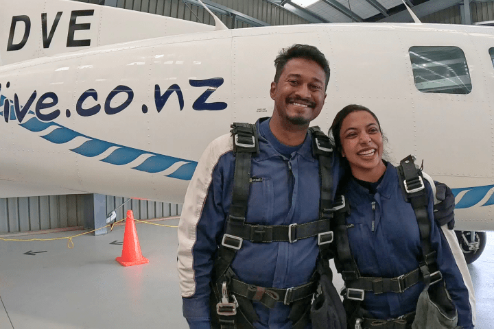 A smiling pair in skydiving gear standing in front of a plane in a hangar.