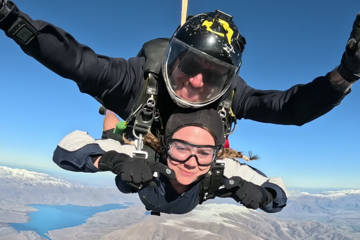 Two people tandem skydiving over mountains and lake under a clear blue sky.