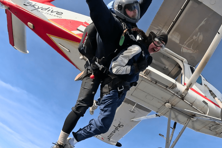 Tandem skydivers jump from a red and white plane against a clear blue sky.