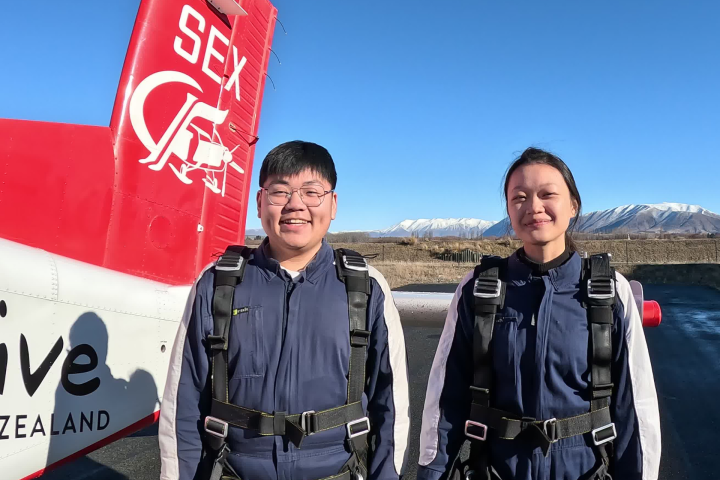 Two people in skydiving suits stand next to a plane with mountains in the background.