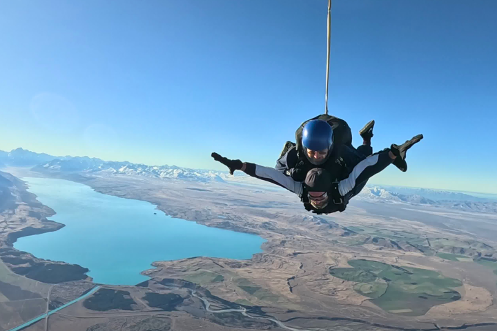 Person skydiving over a lake and mountains under clear blue sky.