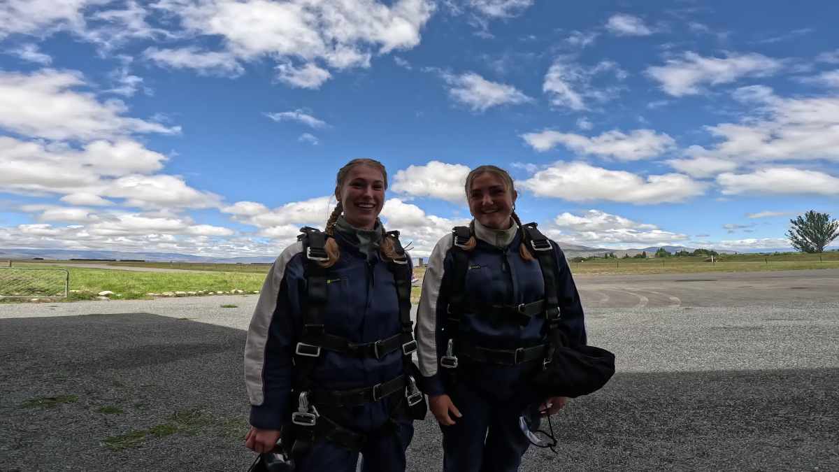 Two people in skydiving gear smiling against a backdrop of sky and grass fields.