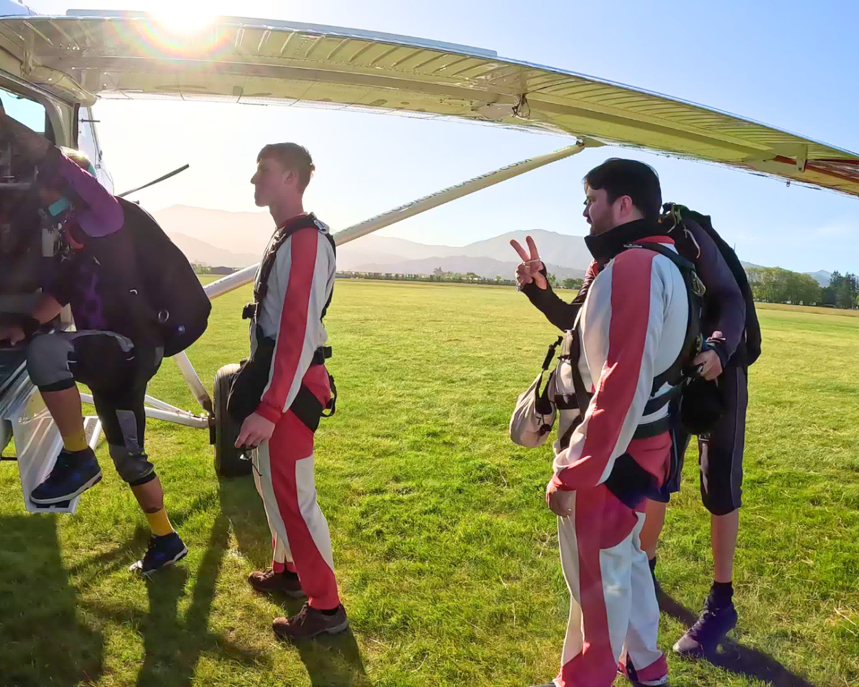 Skydivers in jumpsuits preparing to board a plane, with one raising a peace sign, on a sunny day.
