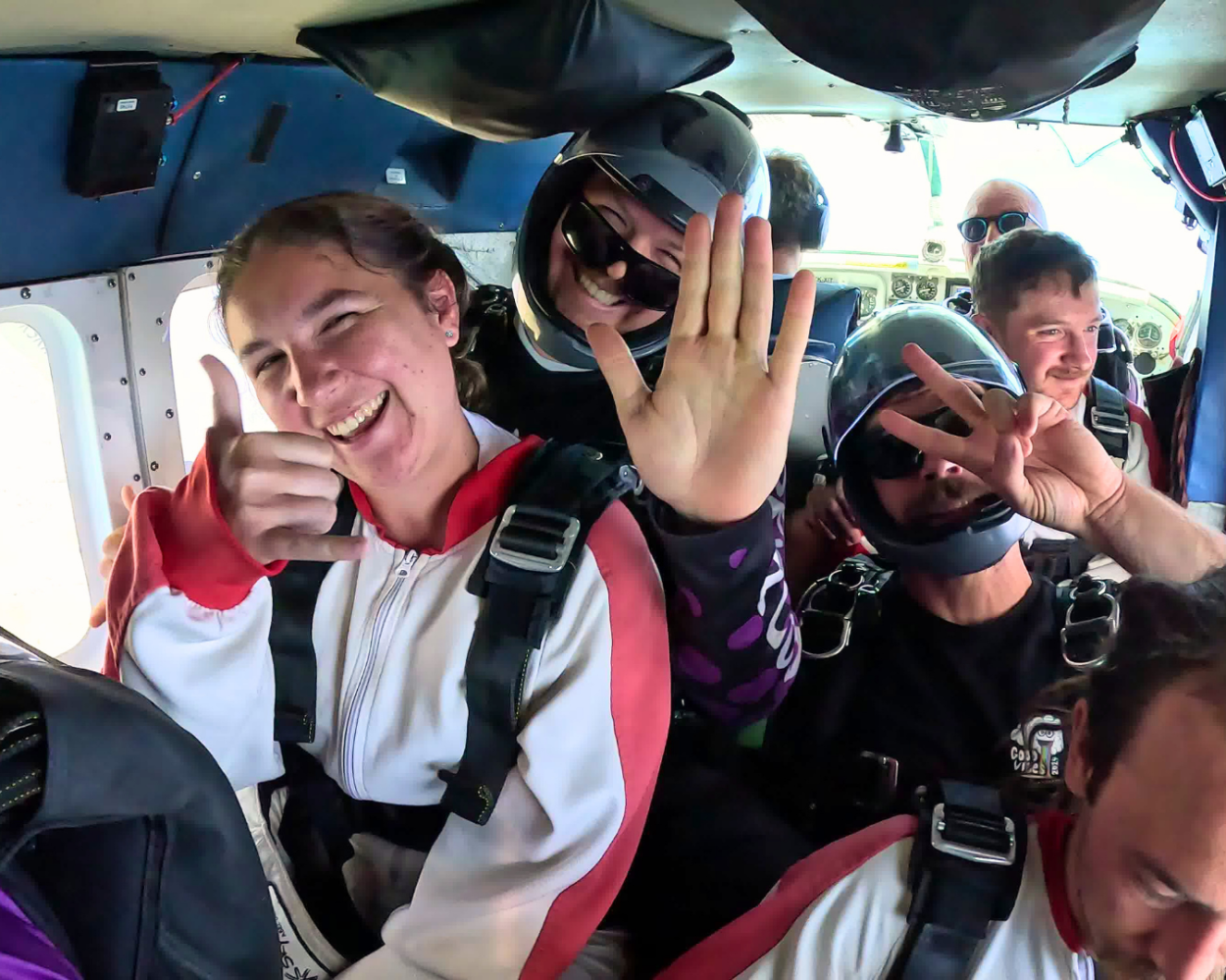 Group of people in skydiving gear smiling inside an airplane.
