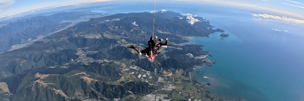 Skydiver tandem in freefall above mountainous coastline with ocean view.