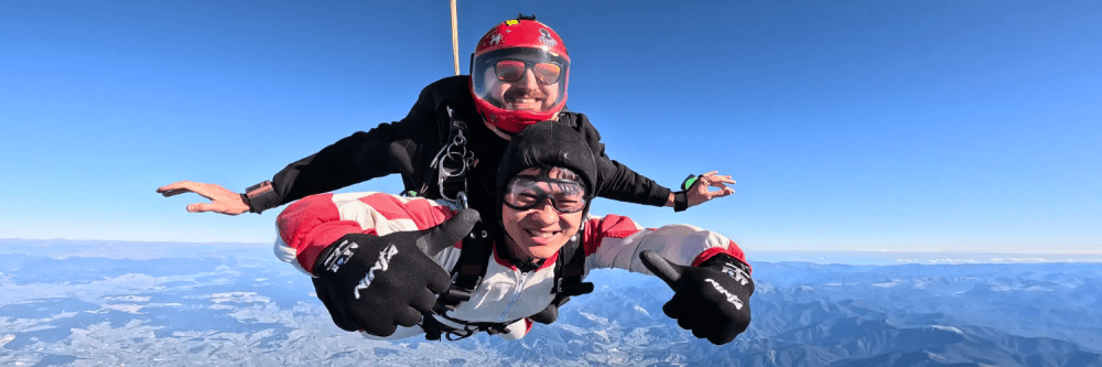 Two people tandem skydiving in clear blue sky over mountainous landscape.