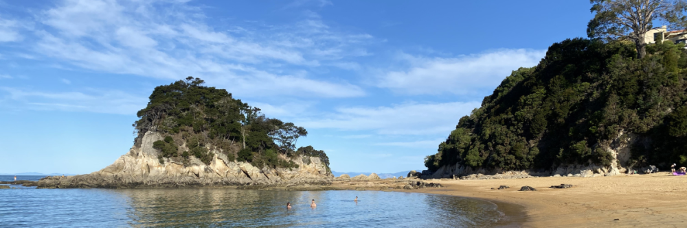 Sunny beach with rocky islands and trees, people swimming in calm water.