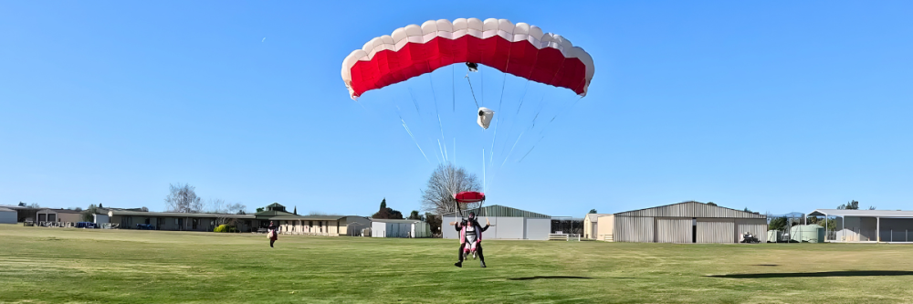 Parachutist landing on grass near buildings under clear blue sky.