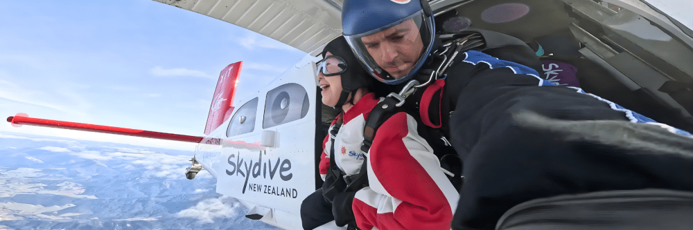 Two people tandem skydiving from a plane labeled 'Skydive New Zealand' above a scenic landscape.