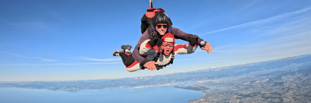 Two people tandem skydiving over a coastal landscape with a clear blue sky.
