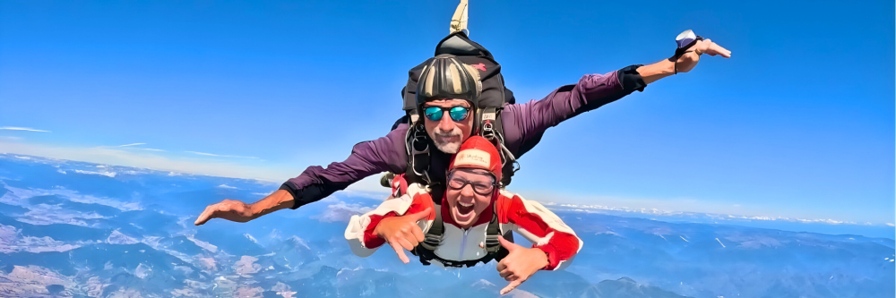 Two people tandem skydiving in a clear blue sky over a mountainous landscape.