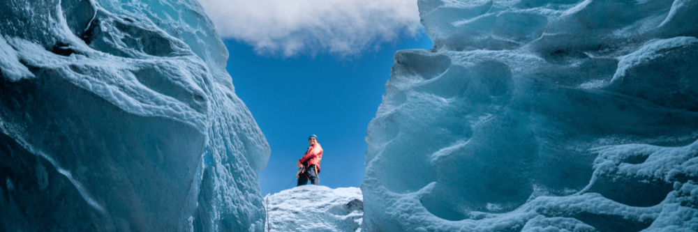 Person in orange jacket standing between large ice formations under blue sky.