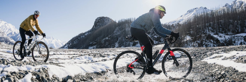 Two cyclists ride on a snowy mountain path with clear skies.