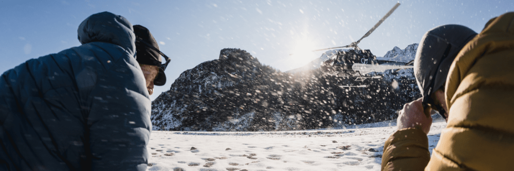 Two people in winter gear watch a helicopter in a snowy mountain landscape.
