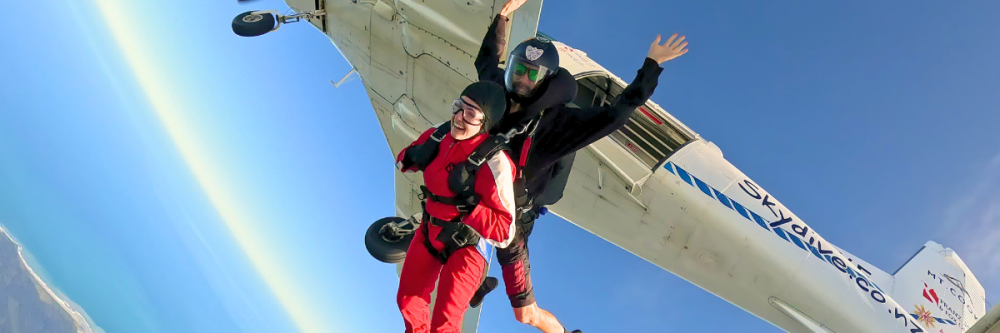 Two people tandem skydiving near an airplane, with ocean and coastline below.