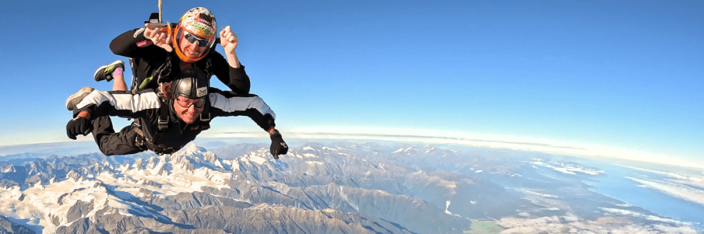 Two people skydiving over a mountainous landscape under a clear blue sky.