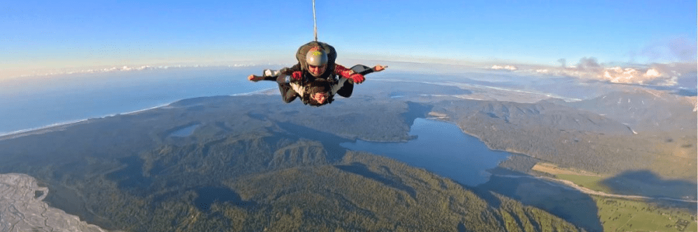 Skydiver soaring over a scenic landscape with forests, mountains, and a lake.