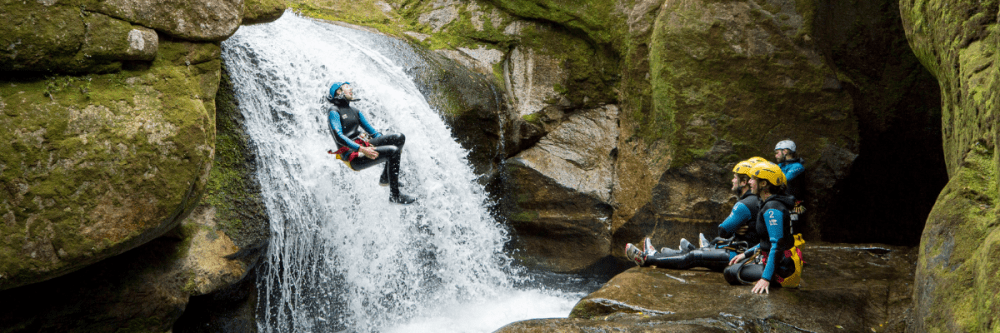 Person sliding down a waterfall while canyoning, others watch in rocky canyon setting.