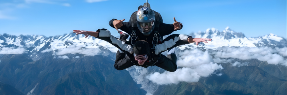 Two people tandem skydiving over snowy mountains and clouds.