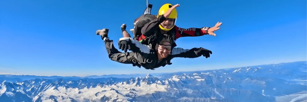Two people tandem skydiving over snow-capped mountains under a clear blue sky.