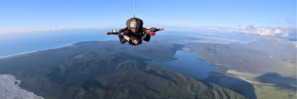 Skydiver descending over scenic landscape with coastline and forests.