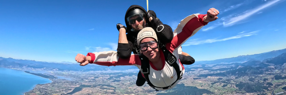 Two people tandem skydiving against a clear blue sky with a landscape view below.