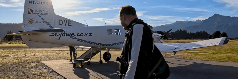 Person in parachute harness walking towards a small plane on an airfield.