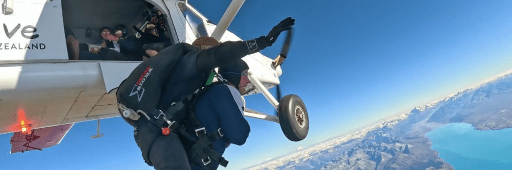 Skydivers exit plane over scenic landscape with mountains and lake.
