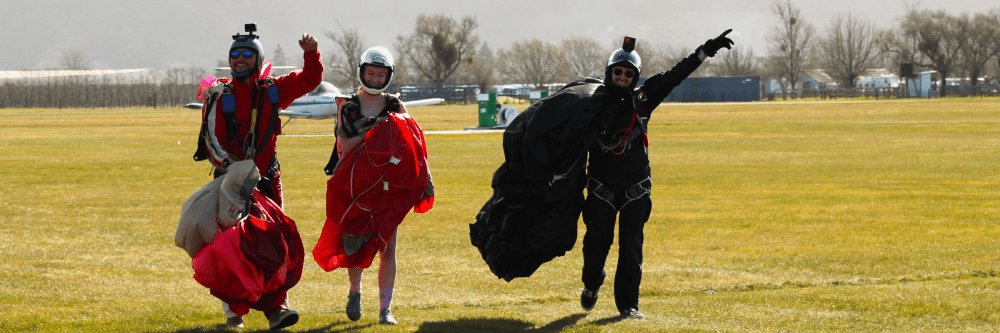 Three skydivers walk on grass carrying parachutes, smiling and pointing.