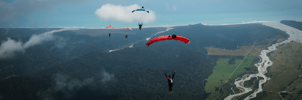 Five skydivers with colorful parachutes above lush landscape and rivers.