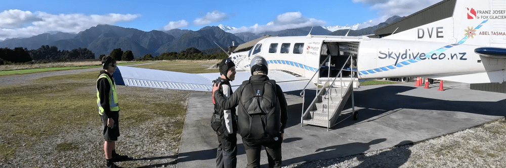 Skydivers boarding a small plane with mountain backdrop.