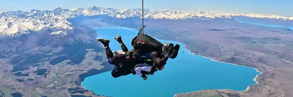 Two people tandem skydiving over a lake with snow-capped mountains in the background.