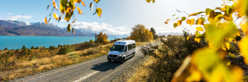 Van on a scenic road beside a lake and mountains, surrounded by autumn trees.