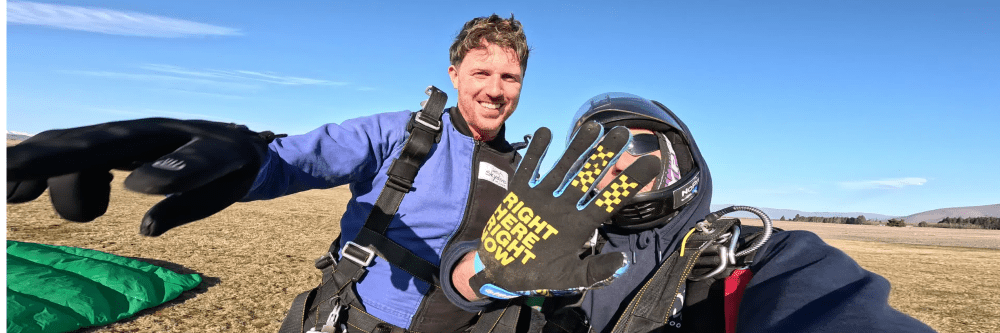 Man and instructor in skydiving gear celebrate on field, smiling, with parachute on ground.