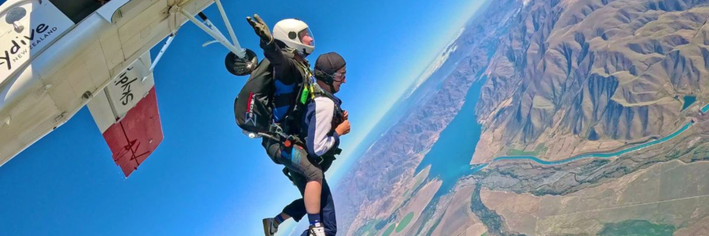 Two people tandem skydiving over a landscape with mountains and a lake.