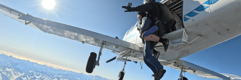 Two people tandem skydiving from a small airplane over snowy mountains.