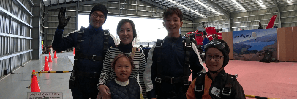 Family in skydiving gear inside a hangar, with a child wearing a helmet and orange suit.