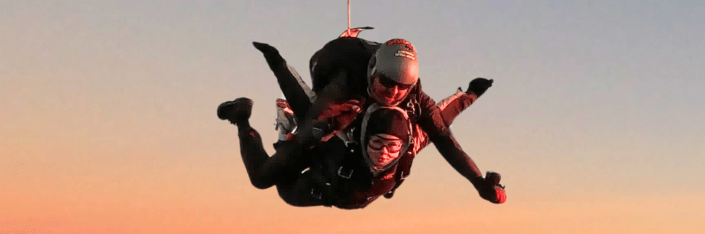 Two people skydiving together, one in front of the other, at sunset.