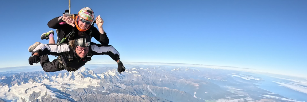 Two skydivers in tandem above mountains and clouds under clear blue sky.